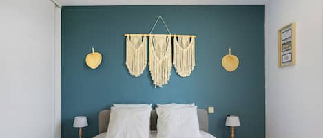 A bedroom with a bed, two bedside tables with lamps, a macrame wall hanging, and a framed picture on the wall.