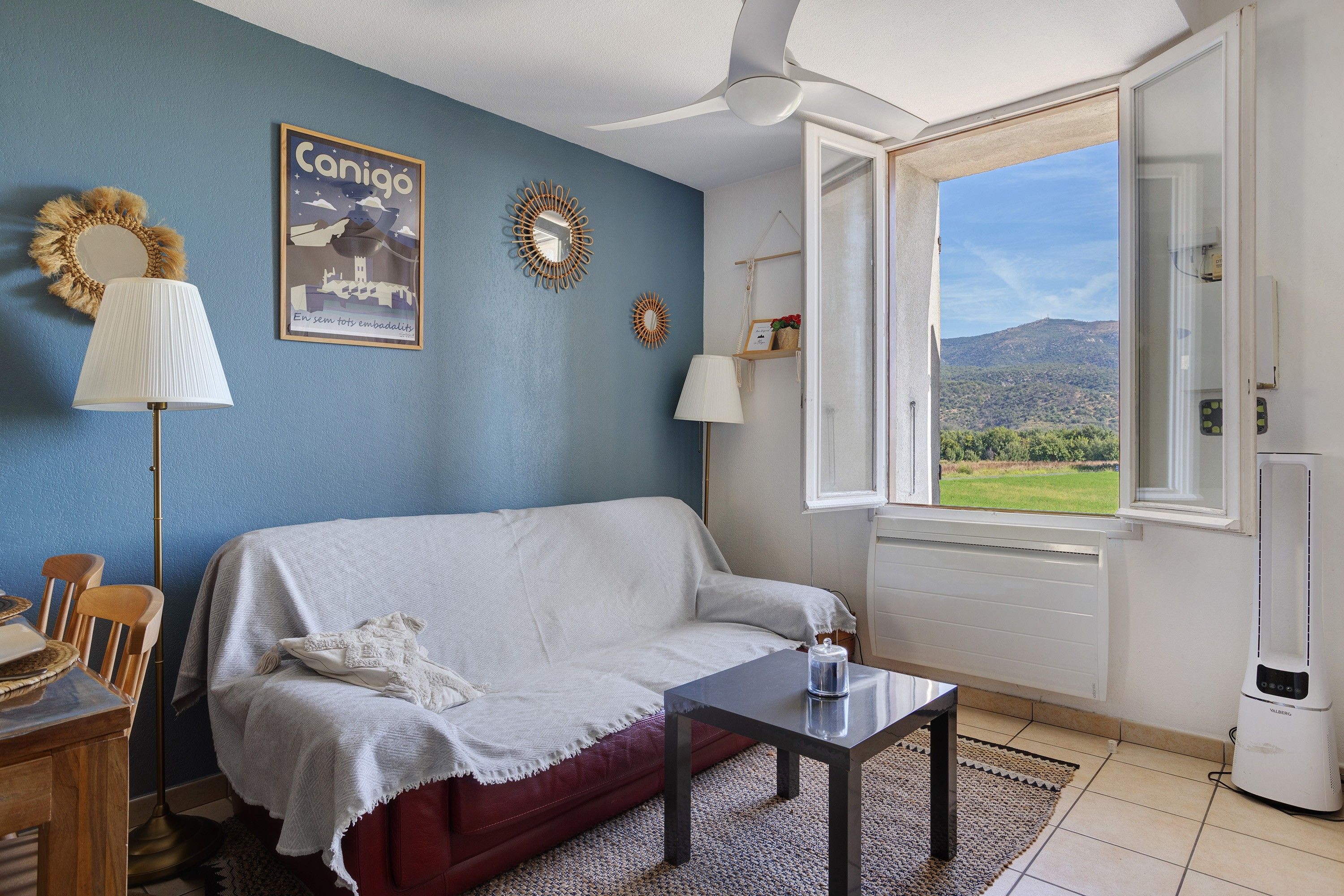 Una habitación con pared azul, sofá blanco, una mesita, una lámpara, un radiador y una ventana con vistas al campo.