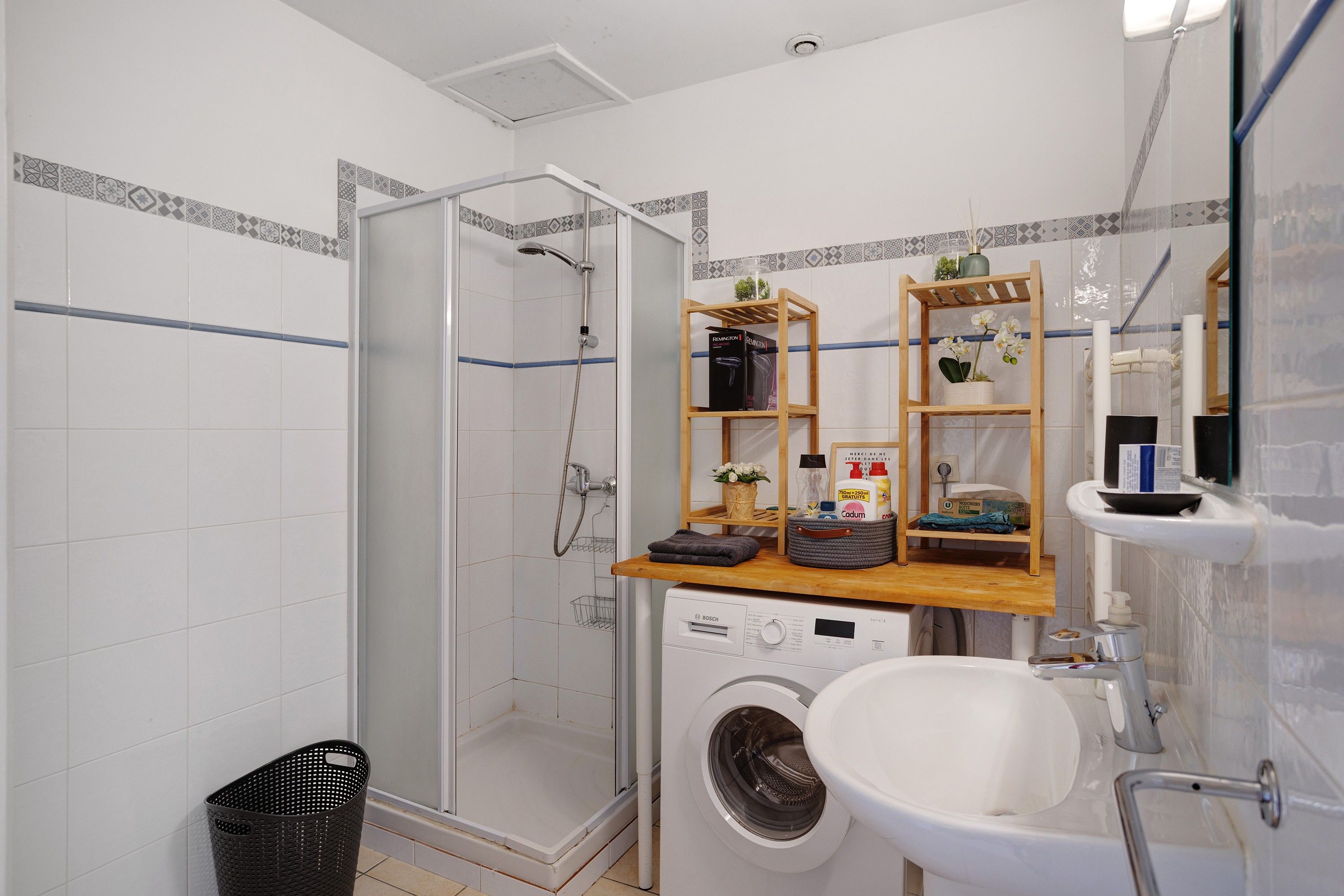 Een badkamer met een douche met glazen wand, een wasmachine, een wastafel en houten planken met toiletartikelen.