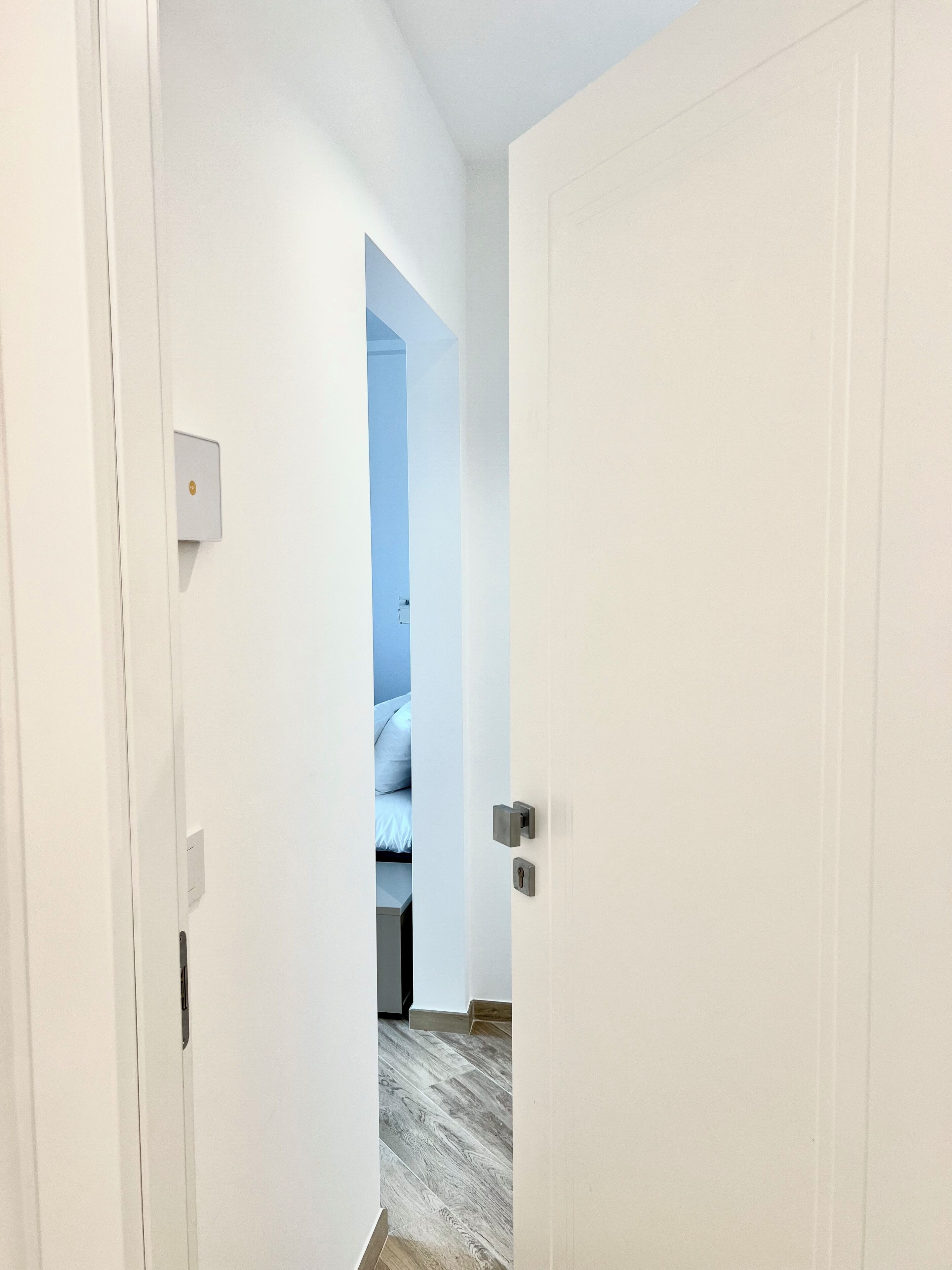 Un couloir avec une porte blanche donnant sur une chambre à coucher, où se trouvent un lit et une table de chevet.