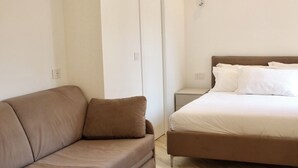 Quarto moderno com sofá marrom, cama com roupa de cama branca e piso de madeira.
