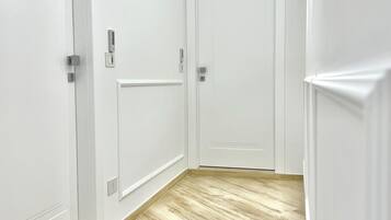 A hallway with white walls and wooden flooring, featuring two white doors with electronic locks and a security camera on the ceiling.