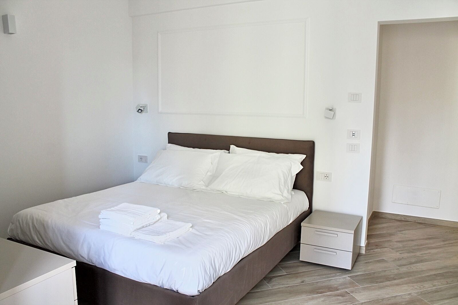 Un lit bien fait, avec du linge de lit blanc et deux oreillers, une table de chevet avec une lampe et un plancher en bois.