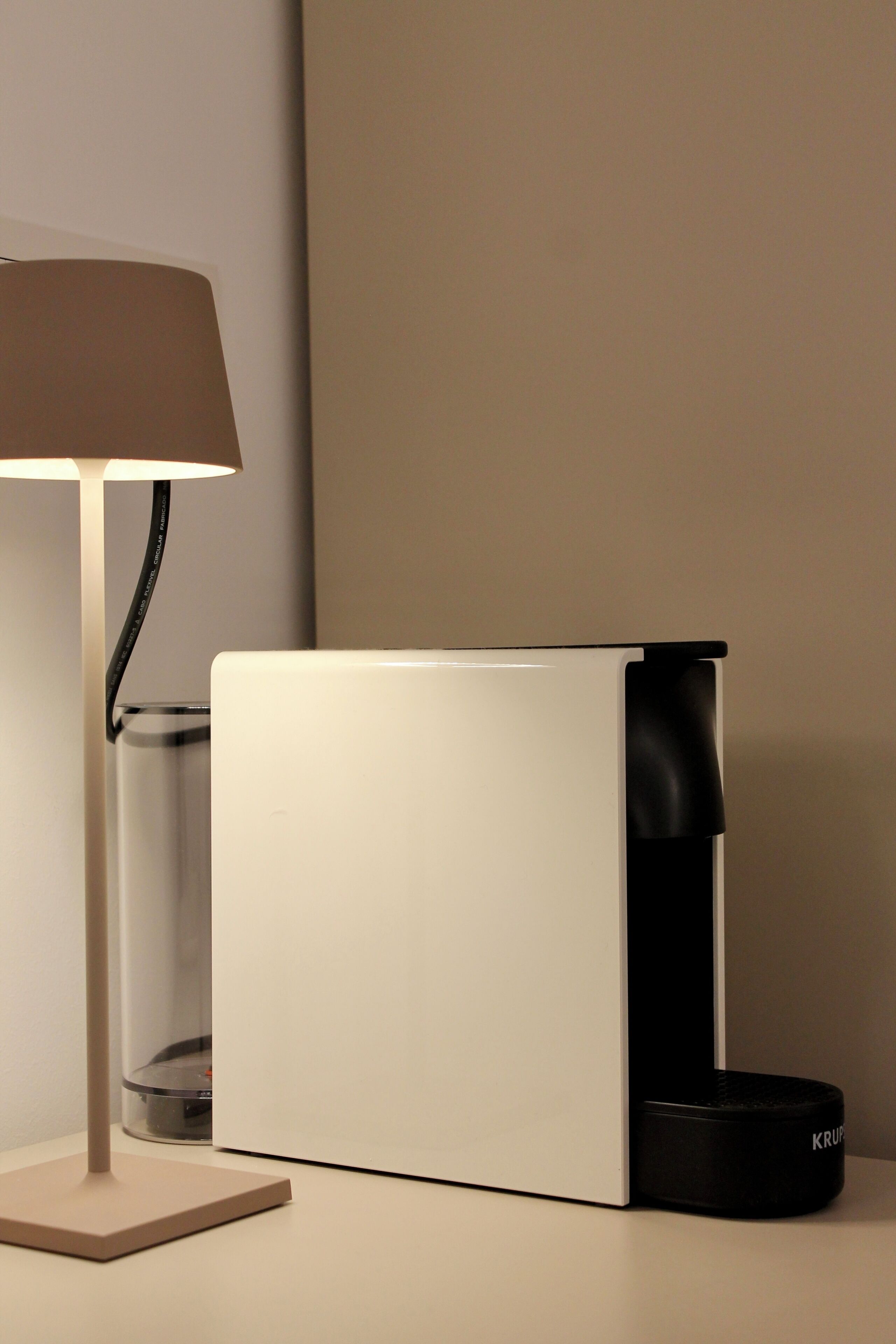 Een moderne lamp met een beige lampenkap en een cilindrische voet, geplaatst naast een wit rechthoekig object met een zwarte, cilindervormige onderdelen.