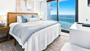 A hotel room with a large bed, a bedside table with a lamp, a nightstand, a bathroom counter, and a balcony with a view of the ocean.