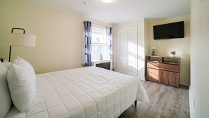Ein Schlafzimmer mit Bett, Nachttisch, Kommode, einem an der Wand befestigten Fernseher und einem Fenster mit Vorhängen.