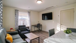 Ein modernes Wohnzimmer mit einer Couch, einem Couchtisch, einem Esstisch mit Pflanze, einem an der Wand montierten Fernseher und einem Fenster mit gemusterten Vorhängen.