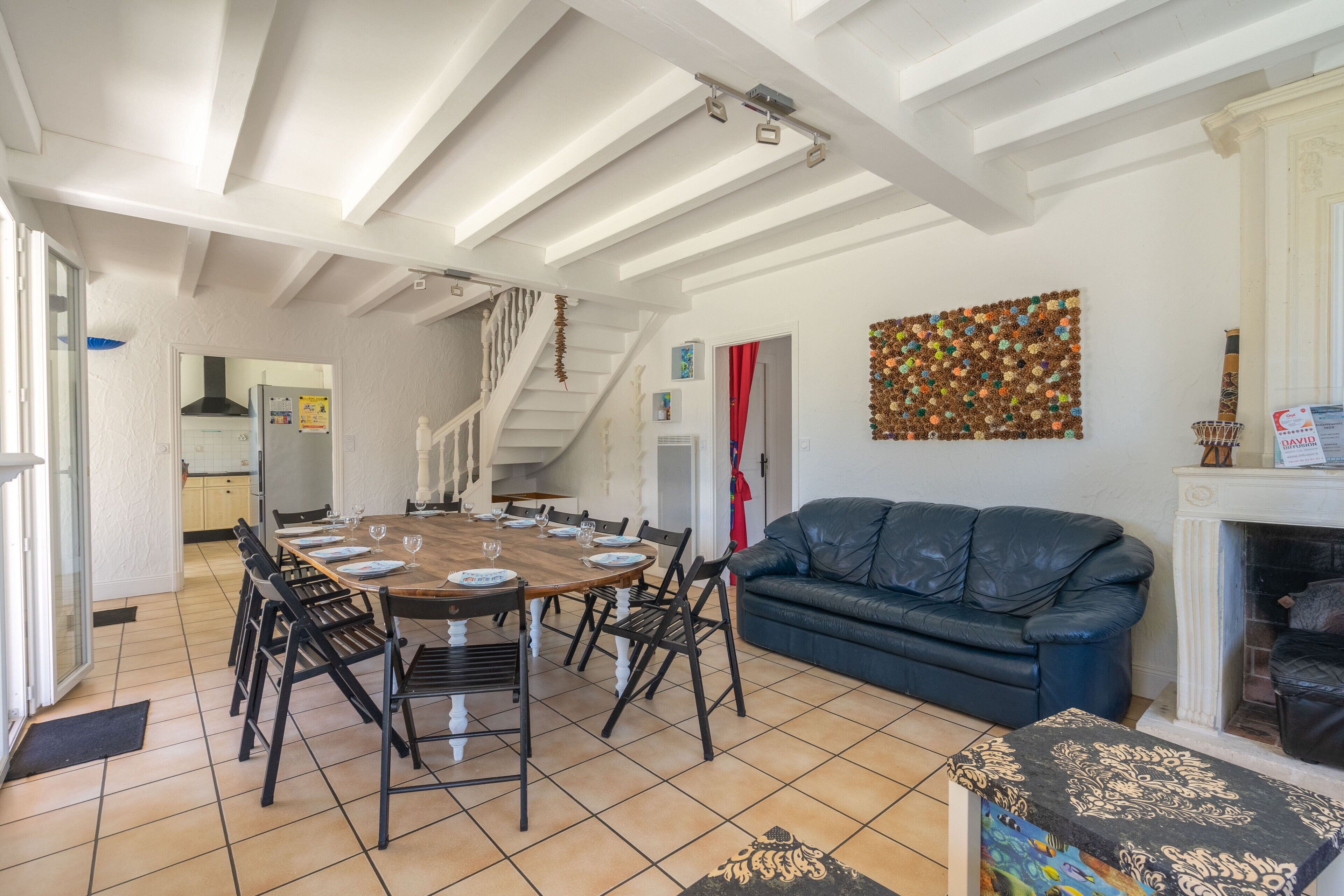 Un espace repas comprenant une table et des chaises en bois, un canapé en cuir noir, une cheminée et une œuvre d’art murale aux couleurs vives.