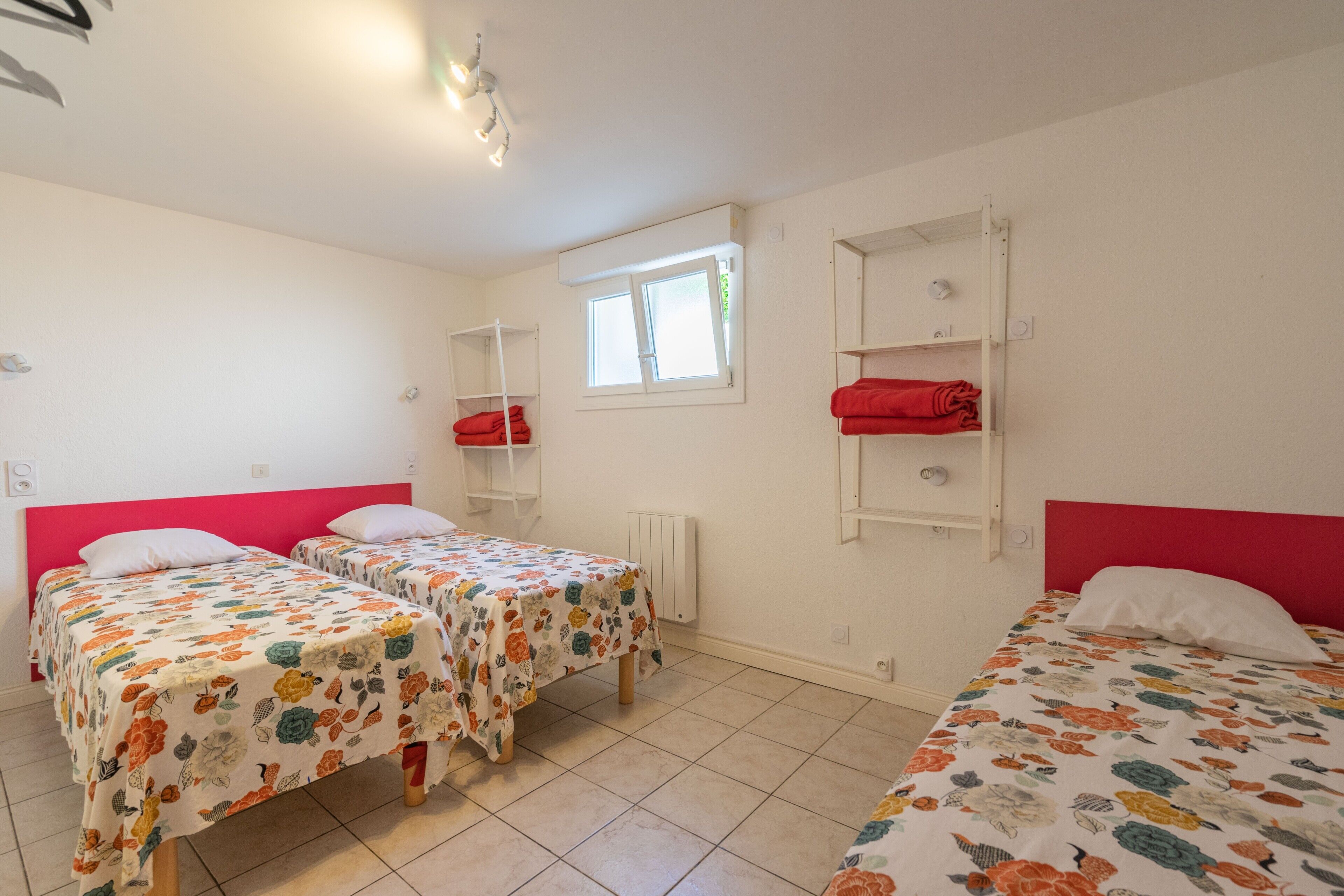 Une pièce avec trois lits, une fenêtre, un radiateur et une étagère sur laquelle se trouve un objet rouge.