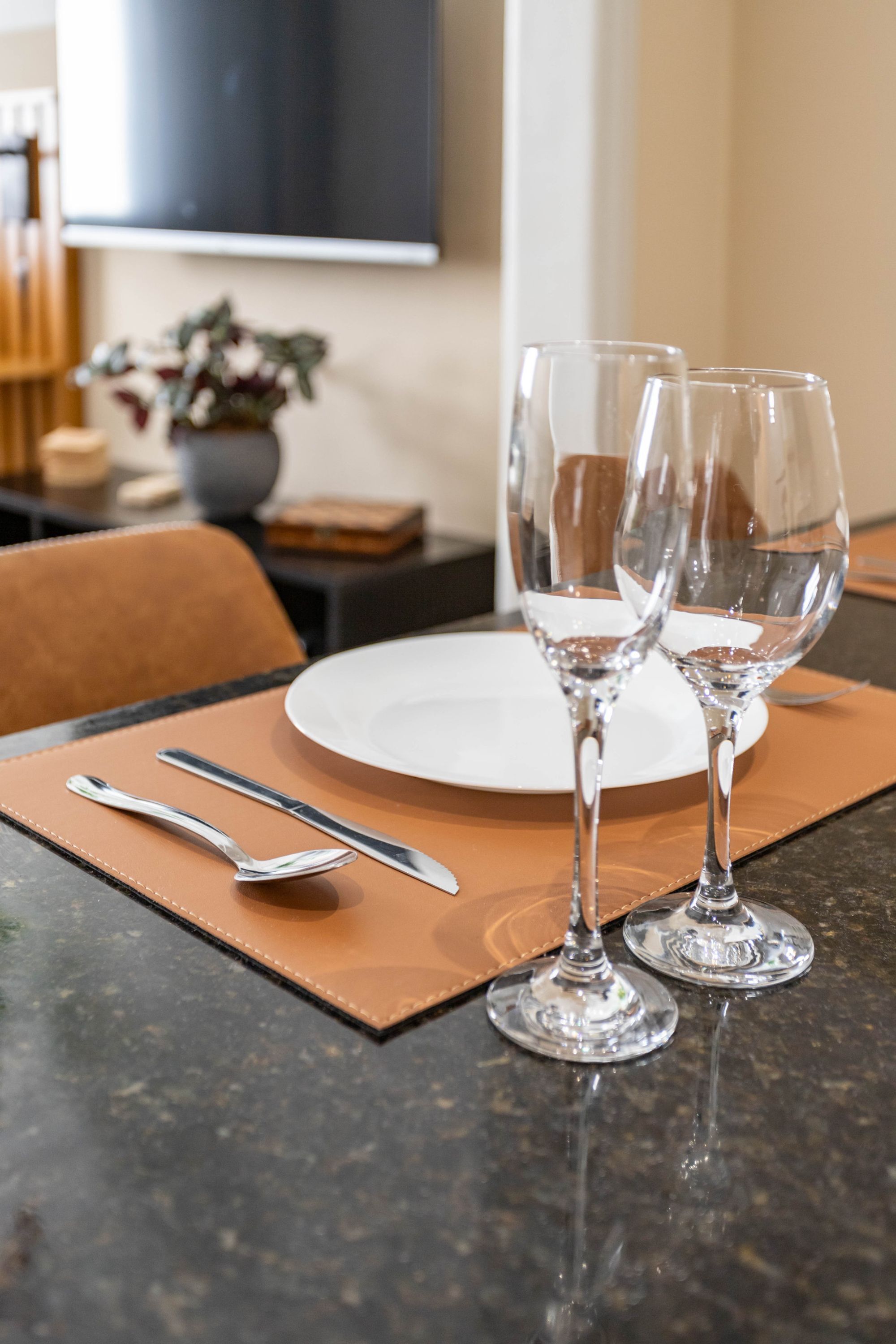 A dining table with a white plate, two wine glasses, and cutlery on a brown placemat.