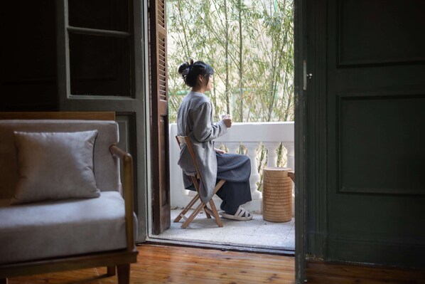 A room with a wooden floor, a sofa, a chair, and a person sitting by a window with a view of bamboo trees.