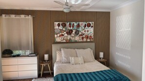 A bedroom with a bed, a ceiling fan, a wall-mounted artwork, a bedside table with a lamp, and a window with curtains. - Seascape Retreat: Time to Slow Down (Agnes Water)