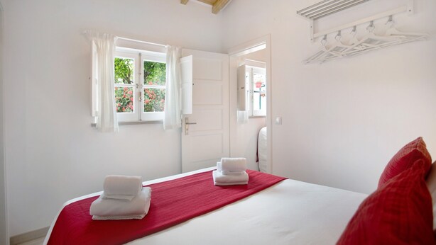 Habitación de hotel con cama, colcha roja, toallas blancas, ventana con cortinas, espejo y perchero.