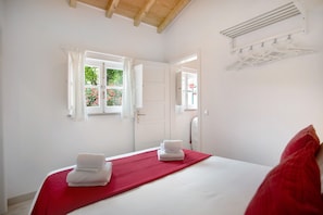 A hotel room with a bed, a red bedspread, white towels, a window with curtains, a mirror, and a coat rack.