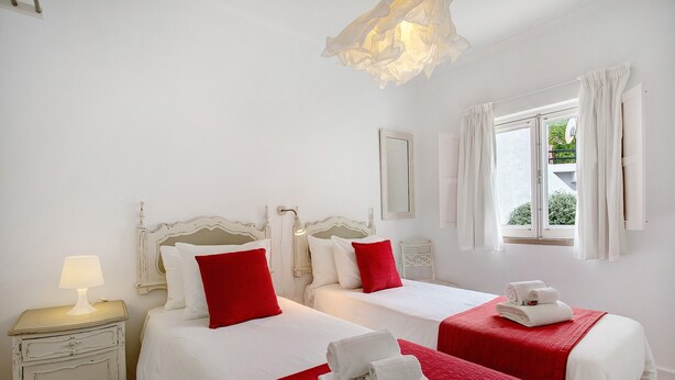 Habitación de hotel con dos camas, ropa de cama blanca, detalles en rojo, una mesita de noche con lámpara y una ventana con cortinas.