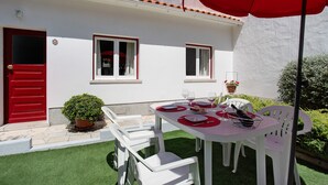 Un edificio blanco con puertas y ventanas rojas, un pequeño jardín con césped artificial, y una mesa para dos con sillas blancas y un paraguas rojo.