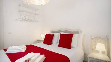 Una habitación de hotel con una cama, ropa de cama roja y blanca, una lámpara blanca sobre una mesita de noche y un estante montado en la pared con una lámpara blanca.