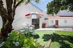 A white building with a red door, a patio with a white table and chairs, and a tree with green leaves.