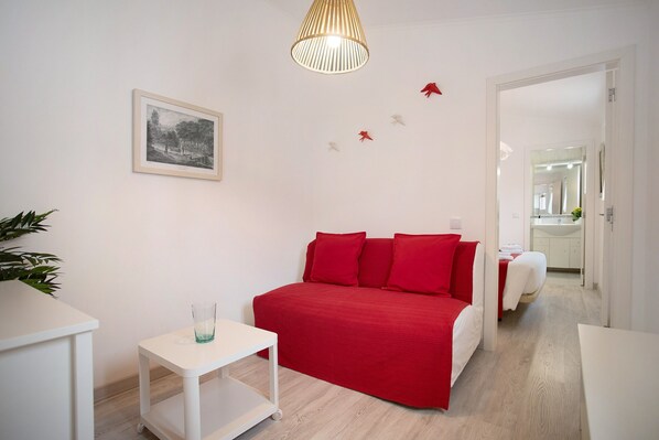 A room with a bed, a small table, a hanging lamp, a framed picture, and a mirror in the background.