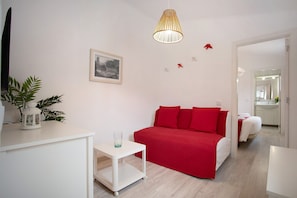 A bedroom with a red bed, white walls, a small white table, a hanging light fixture, and a framed picture on the wall.