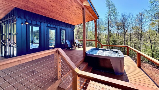 Een houten terras met een hottub, buitenmeubels en uitzicht op een bos.