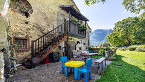 Een rustiek stenen gebouw met een houten trap die leidt naar een balkon, buitenmeubels met blauwe en gele stoelen, en een uitzicht op een weelderig groen landschap met bergen op de achtergrond.