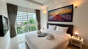 A hotel room with a large bed, a TV, a painting of a sunset, and a view of a building outside. - 6-32 - 1 Bdr Condo With Balcony and Shared Pool (Patong)
