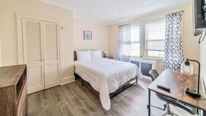 A bedroom with a bed, a desk, a chair, a wardrobe, and a window with patterned curtains. - Renovated 1-Bedroom Apartment in Beautiful Downtown Falmouth Located on Main St (Falmouth)