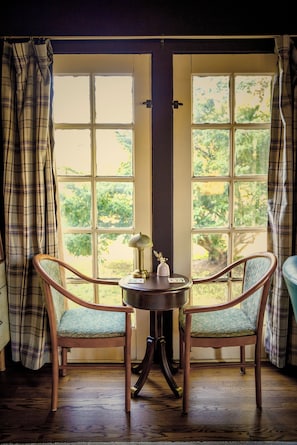 Une pièce dotée d’un plancher en bois, de deux chaises avec des sièges rembourrés, d’une petite table ronde avec une lampe, et offrant une vue à travers de grandes fenêtres ornées de rideaux.
