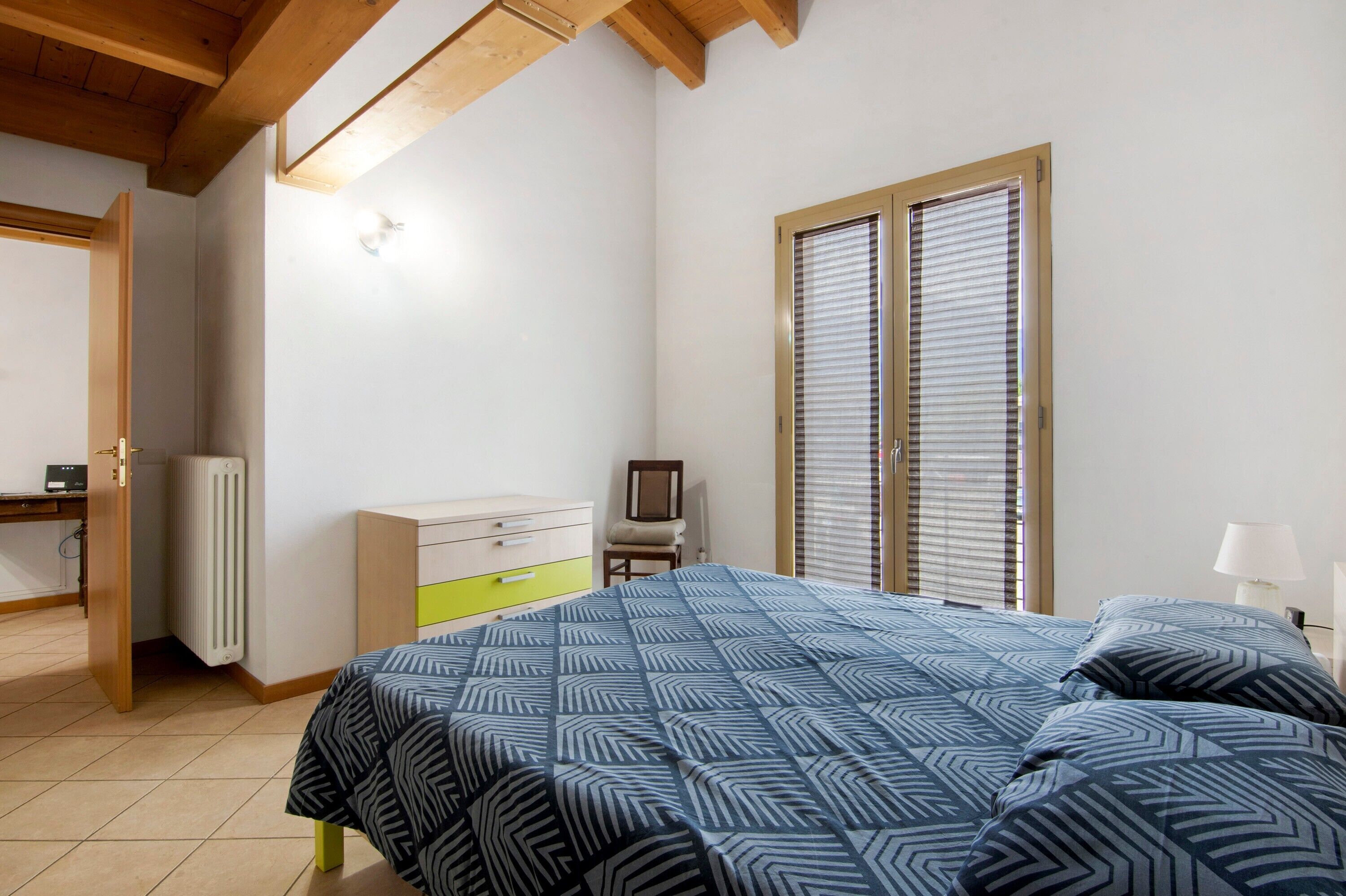 Une chambre à coucher comprenant un lit, une commode, un radiateur, un bureau et une fenêtre avec des stores.