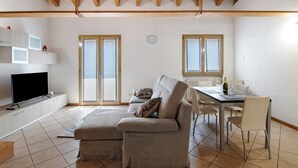 A living room with a sofa, a TV, a dining table with chairs, and a window with blinds. - Apartment 'Casa Zia Dani' with Mountain View, Private Terrace and Wi-Fi (Piantedo)