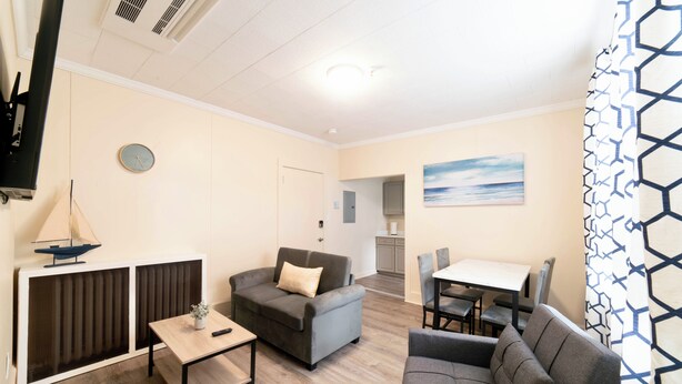 Ein modernes Wohnzimmer mit einer Couch, einem kleinen Tisch und einem Fernseher. An der Wand hängt ein Bild und es gibt ein Fenster mit Vorhängen.