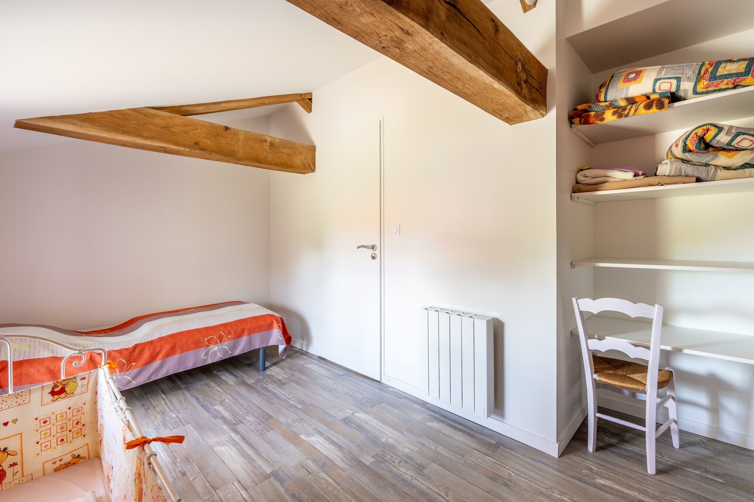 Une pièce comprenant un lit, une chaise, un radiateur et une étagère avec des couvertures pliées.