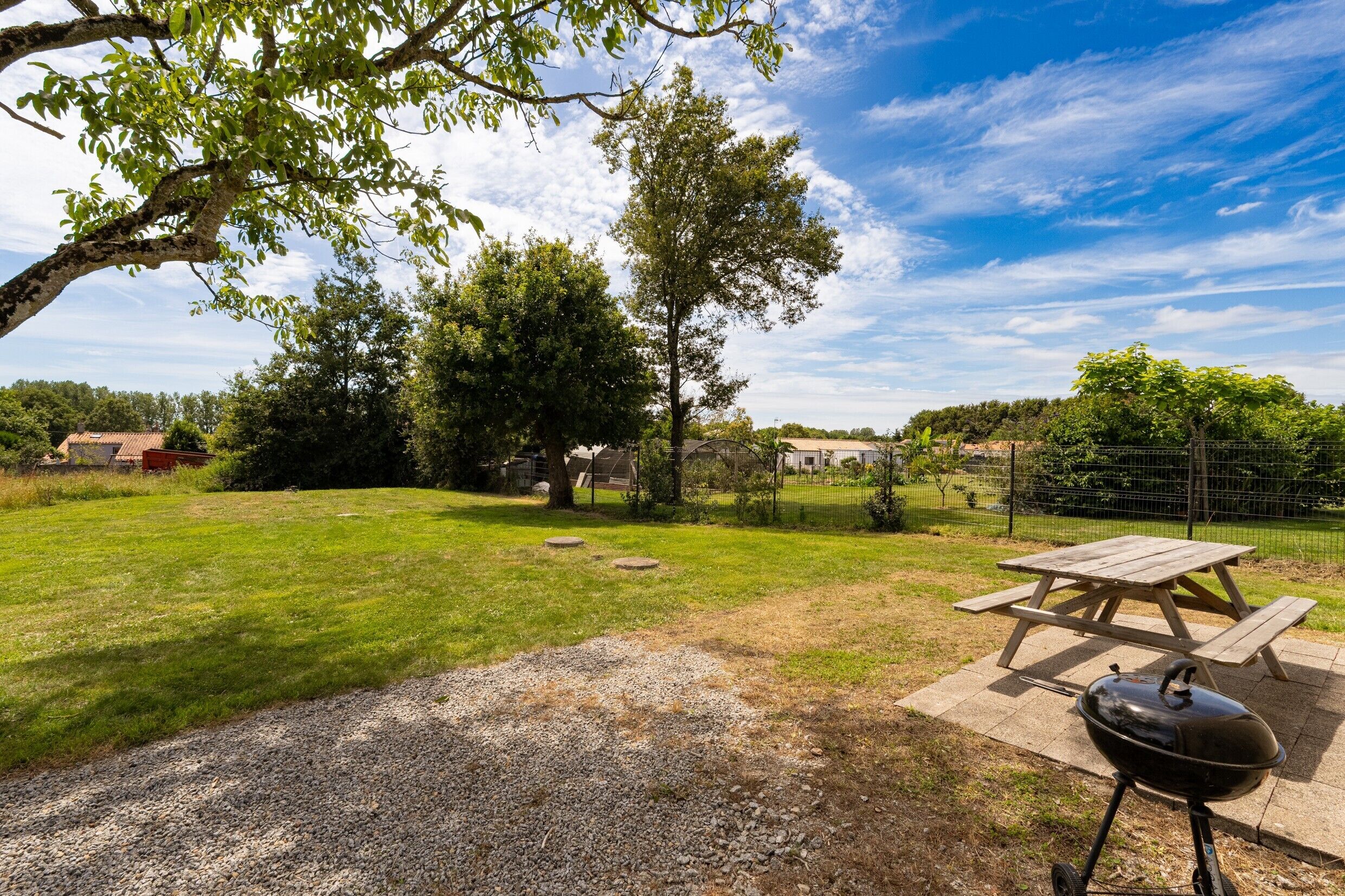 Ein Garten mit Picknicktisch und Grill, umgeben von Bäumen und blauem Himmel.