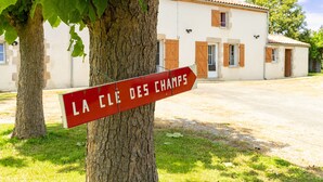 Un cartel con la inscripción 'LA CLE DES CHAMPS', un árbol y un edificio con tejado de tejas al fondo.