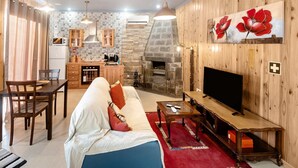 Un salon comprenant un canapé, un téléviseur et une cheminée, attenant à une cuisine avec des meubles en bois et un mur en pierre.