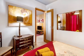 Quarto de hotel com cama, cómoda de madeira, secretária, candeeiro, pintura grande, espelho e porta para outro quarto.