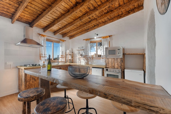 A rustic kitchen with wooden beams, a central island with stools, a microwave, and a refrigerator.