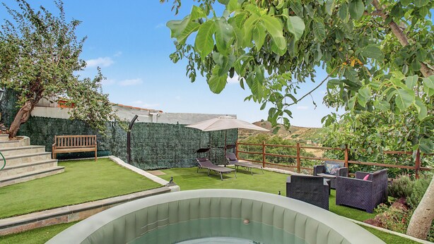 Une piscine extérieure dotée d’une terrasse en bois, d’un banc et d’un jardin clos, agrémenté de diverses plantes et offrant une vue sur la campagne.