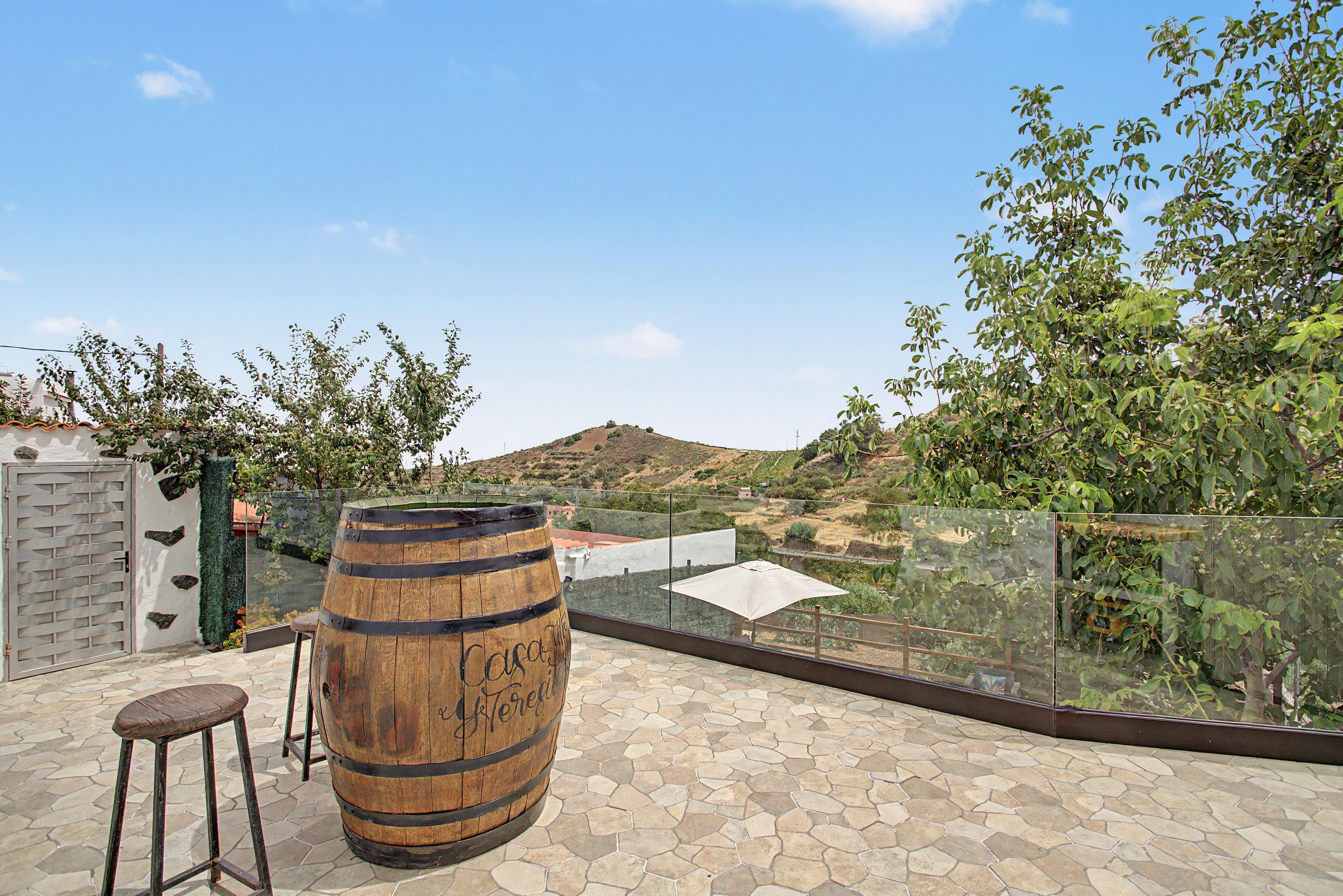 Una terraza en la azotea con un barril de madera, barandilla de cristal y vistas a un paisaje montañoso.
