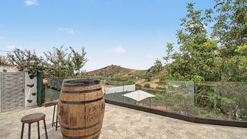 Una terraza en la azotea con un barril de madera, barandilla de cristal y vistas a un paisaje montañoso.