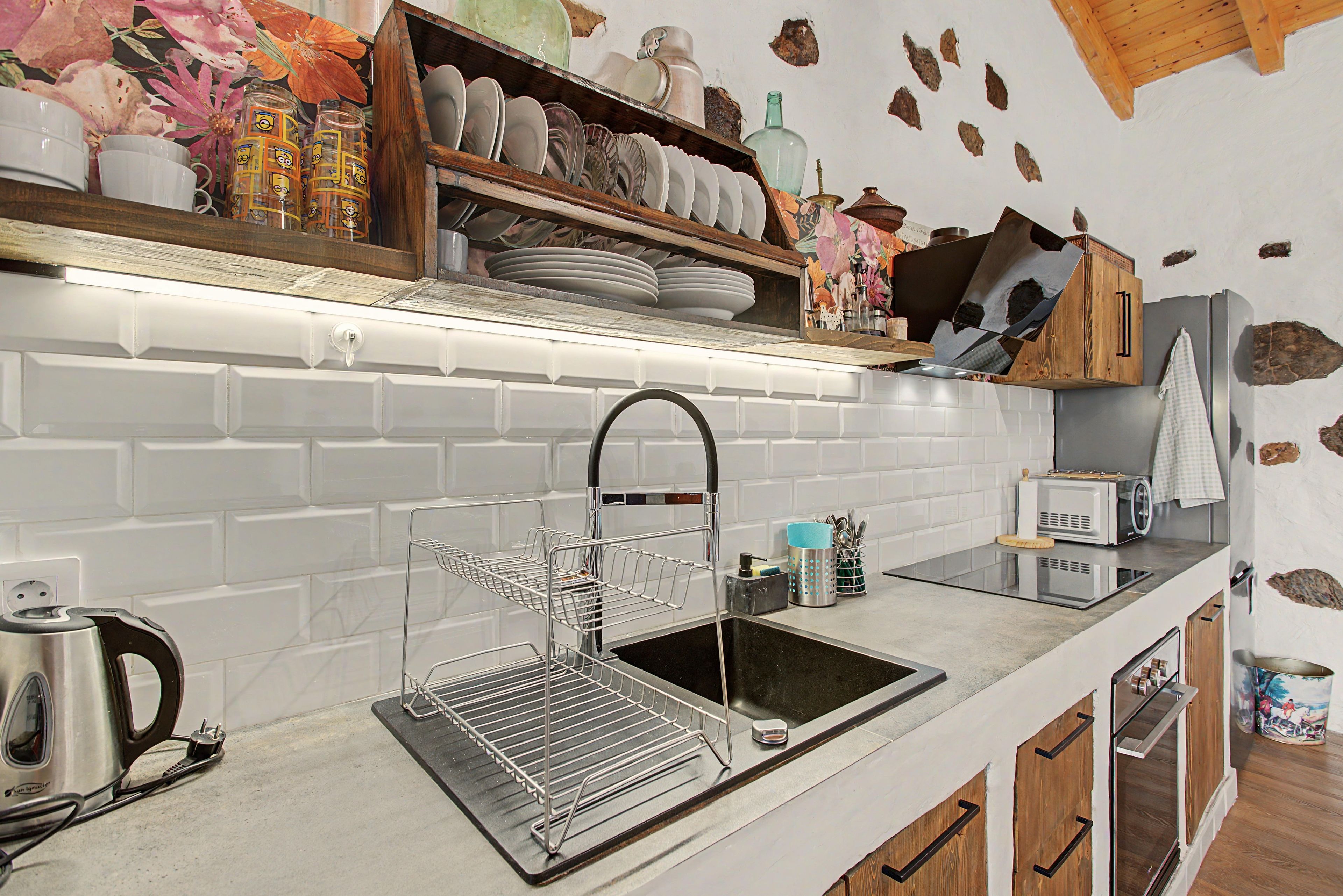 Una cocina con salpicadero blanco de azulejos, un lavamanos de acero inoxidable, un escurreplatos, una tetera y armarios de madera.