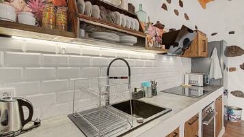 Una cocina con salpicadero blanco de azulejos, un lavamanos de acero inoxidable, un escurreplatos, una tetera y armarios de madera.