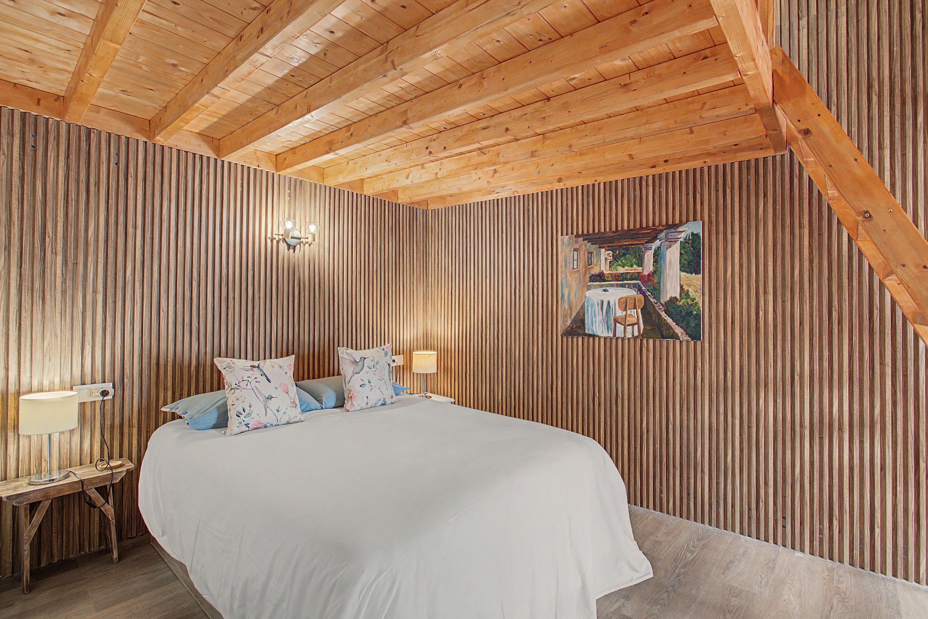 Ein Schlafzimmer mit schrägem Dach, Holzpaneelen, einem Bett mit weißer Bettwäsche, zwei Nachttischlampen und einem Bild an der Wand.