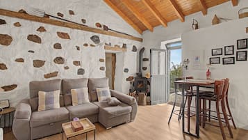 A cozy living room with a stone wall, a wooden ceiling, a sofa, a coffee table, and a dining area with chairs and a table.