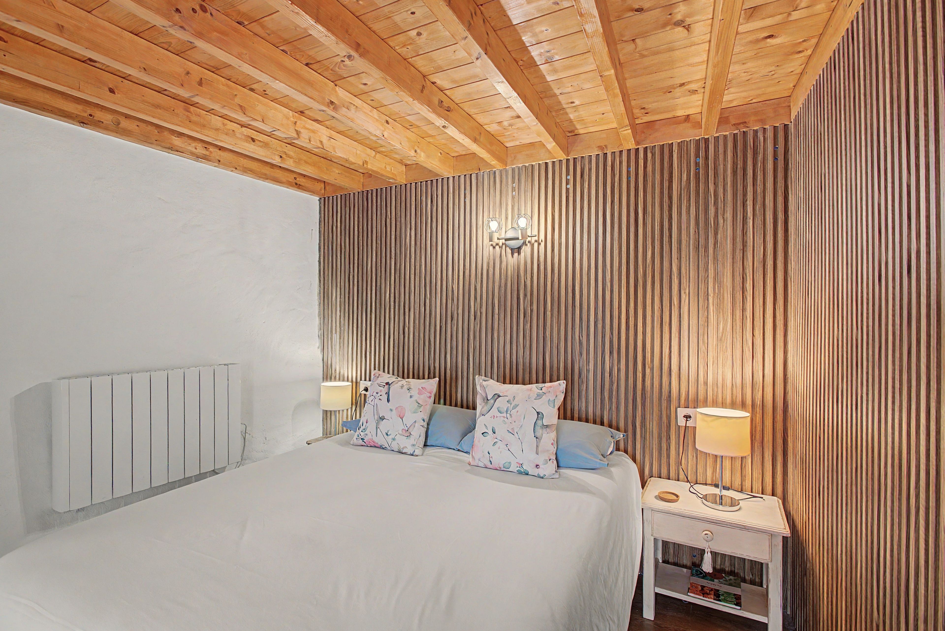 Un dormitorio con techo de madera, una cama con almohadas florales, una mesita de noche con una lámpara y un aplique de pared.