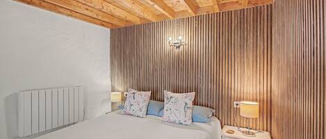 Un dormitorio con techo de madera, una cama con almohadas florales, una mesita de noche con una lámpara y un aplique de pared.