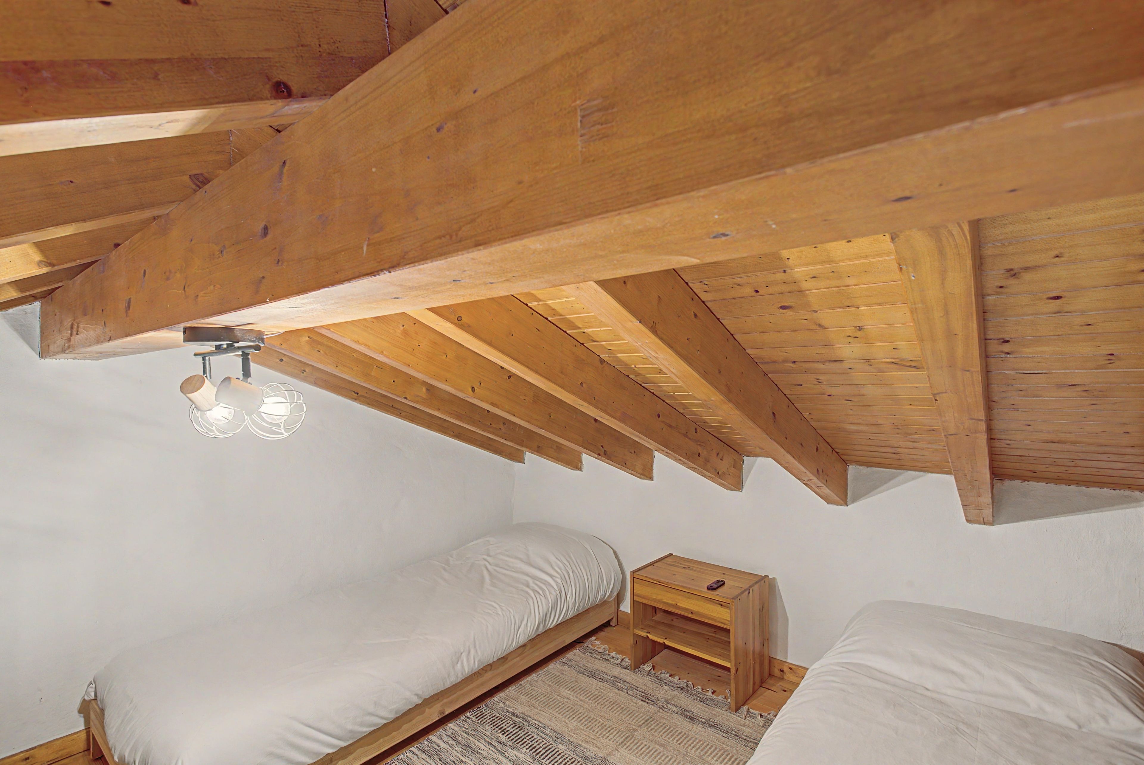 Un dormitorio con techo inclinado, vigas de madera, una cama, una mesita de noche y una lámpara fijada a la pared.