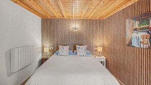 Une chambre avec un plafond en bois, un lit agrémenté d’oreillers à motifs floraux, une table de chevet avec une lampe, et un mur recouvert de lambris en bois verticaux.