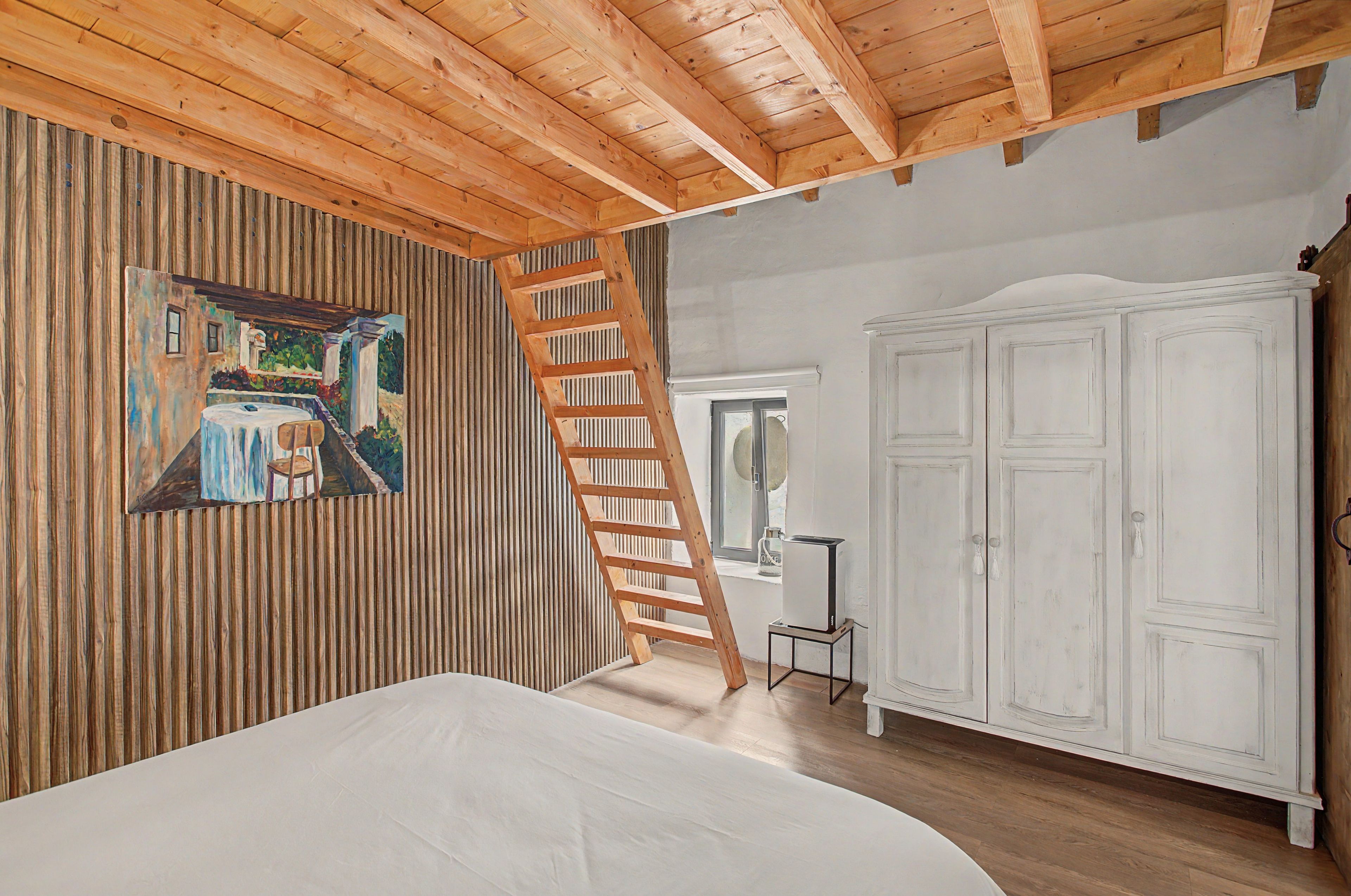 Ein Schlafzimmer mit Holzbalkendecke, einem Bild an der Wand, einer Leiter, einem Kleiderschrank und einem kleinen Tisch.